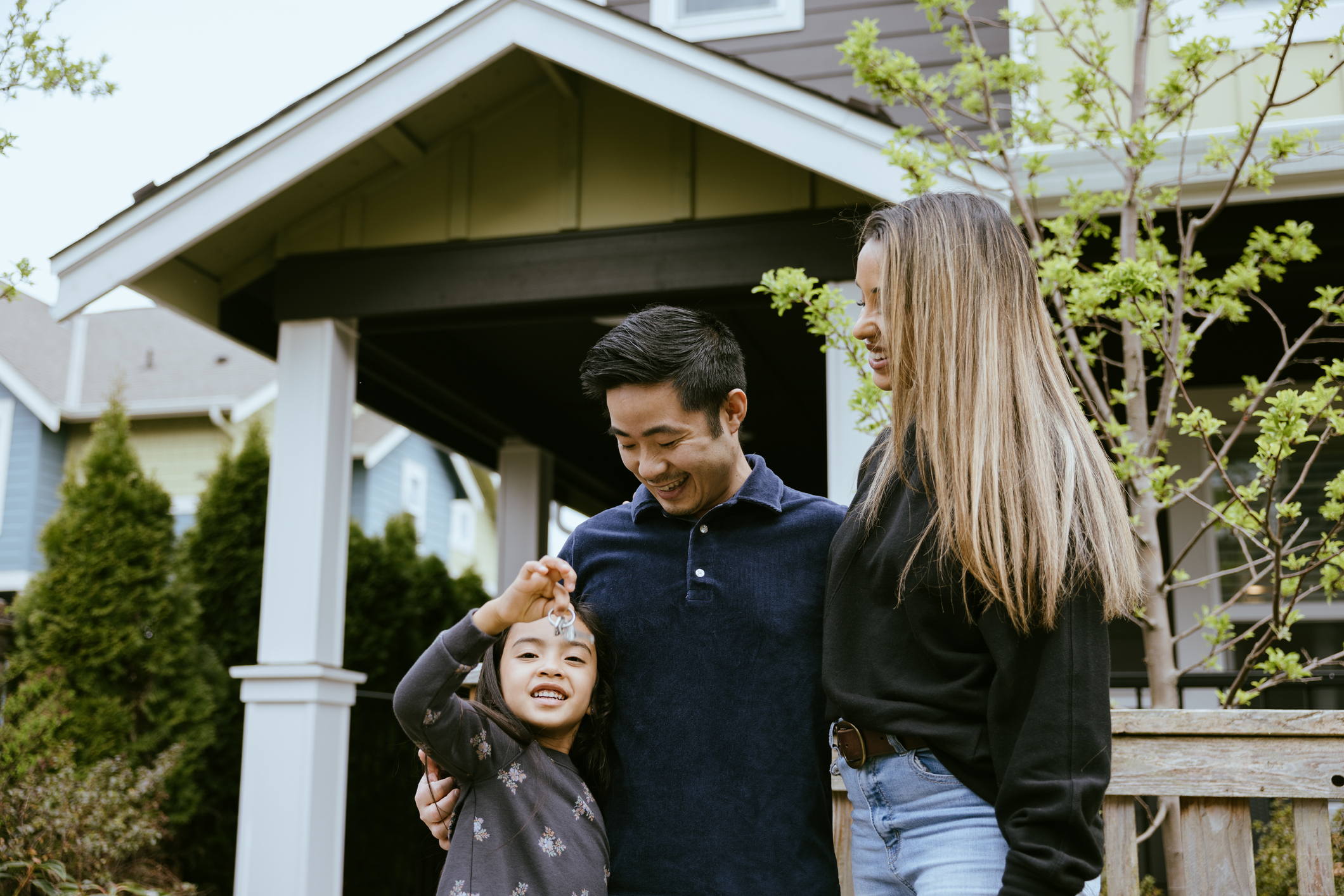 A husband, wife, and their daughter hold up the keys to their new house, smiling with joy and excitement at their investment.