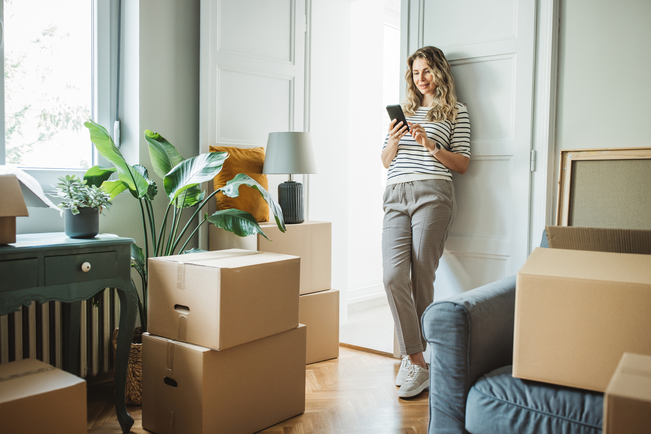 A smiling middle-aged woman is on her smartphone while surrounded by moving boxes and some furniture.