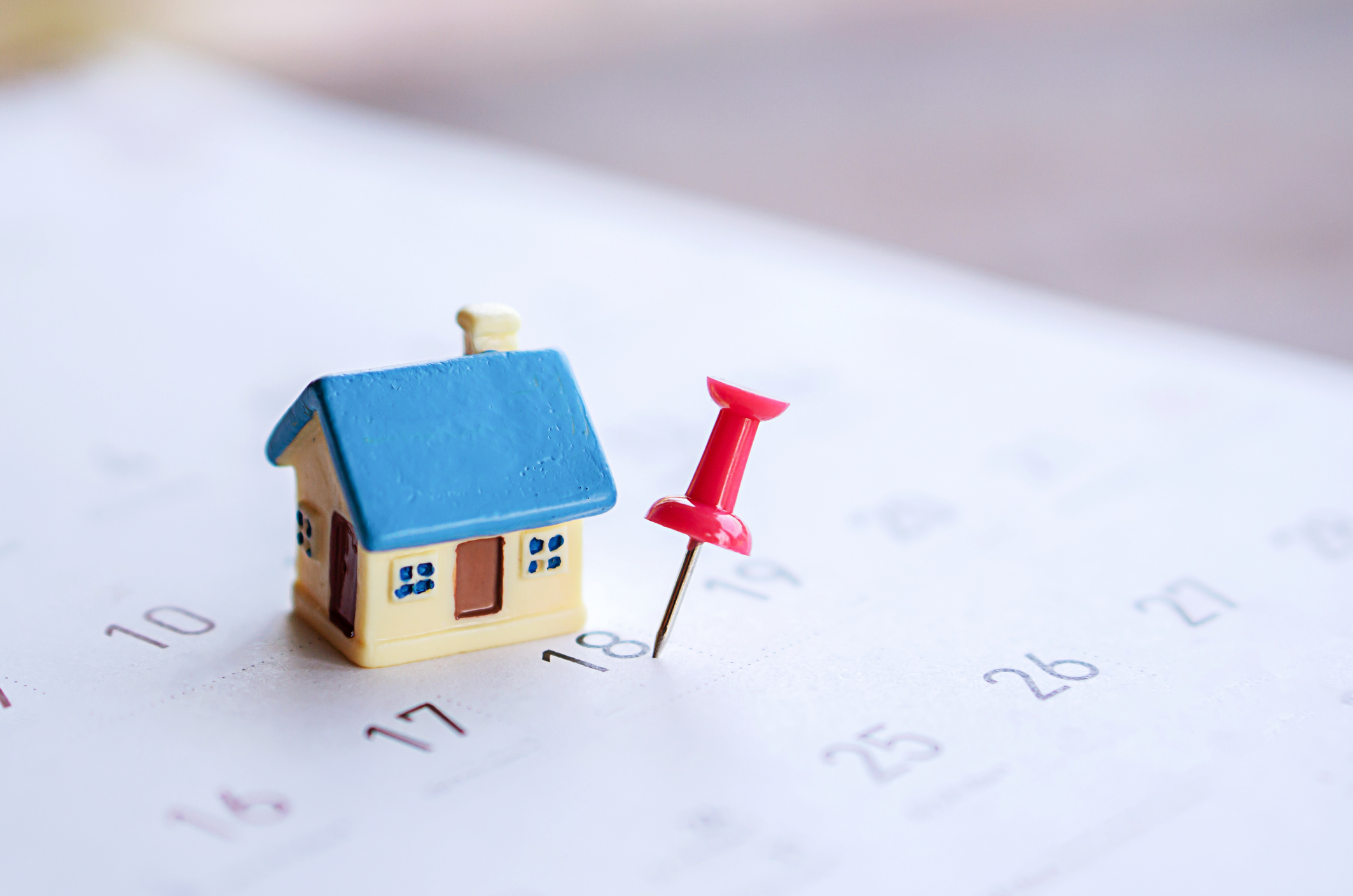 A small house figurine with a blue roof sits on top of a calendar with a red thumbtack stuck on the date of the 18th.