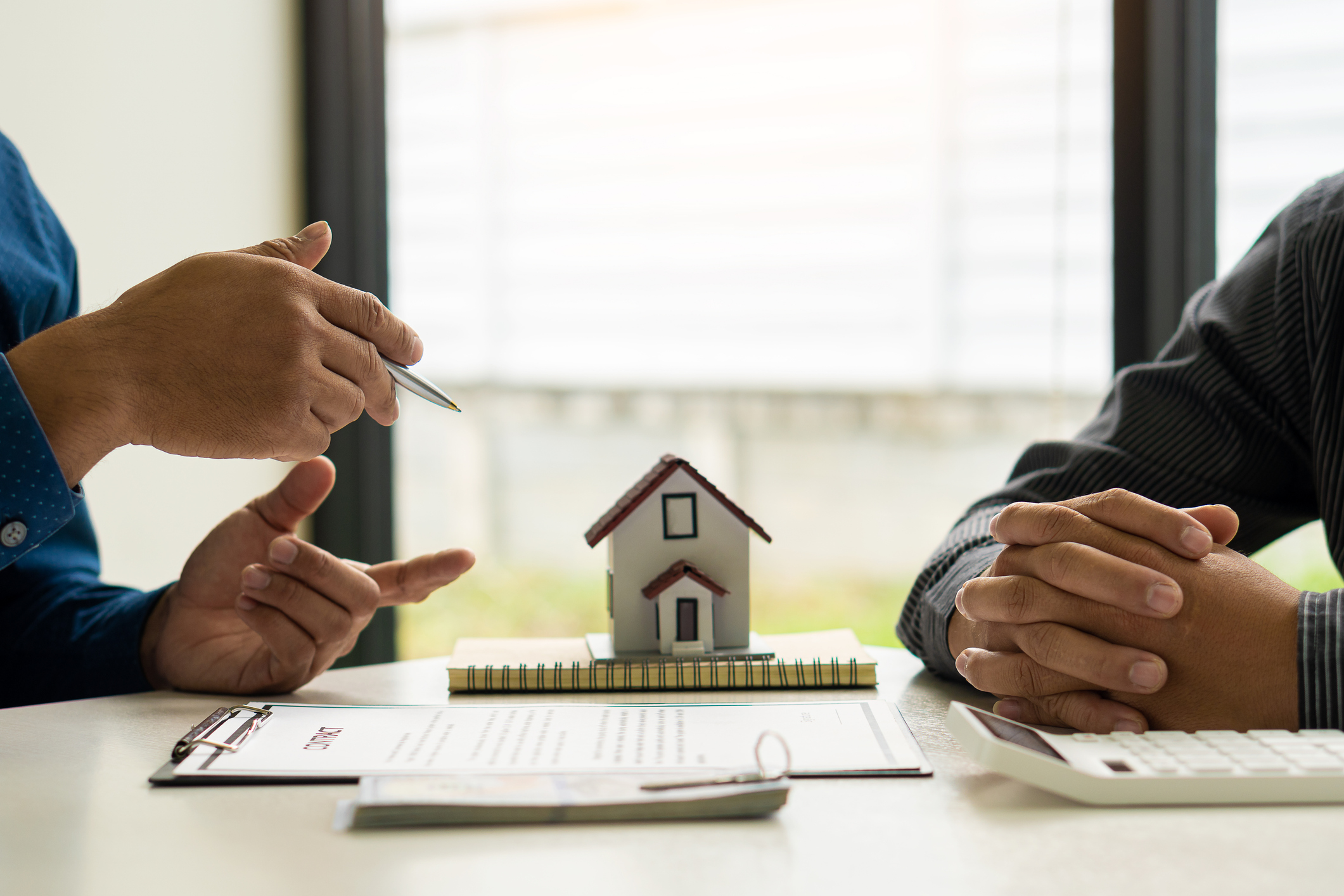 Close-up of the hands of two people in active discussion with a contract laid out on the table, along with a calculator and a small model home.