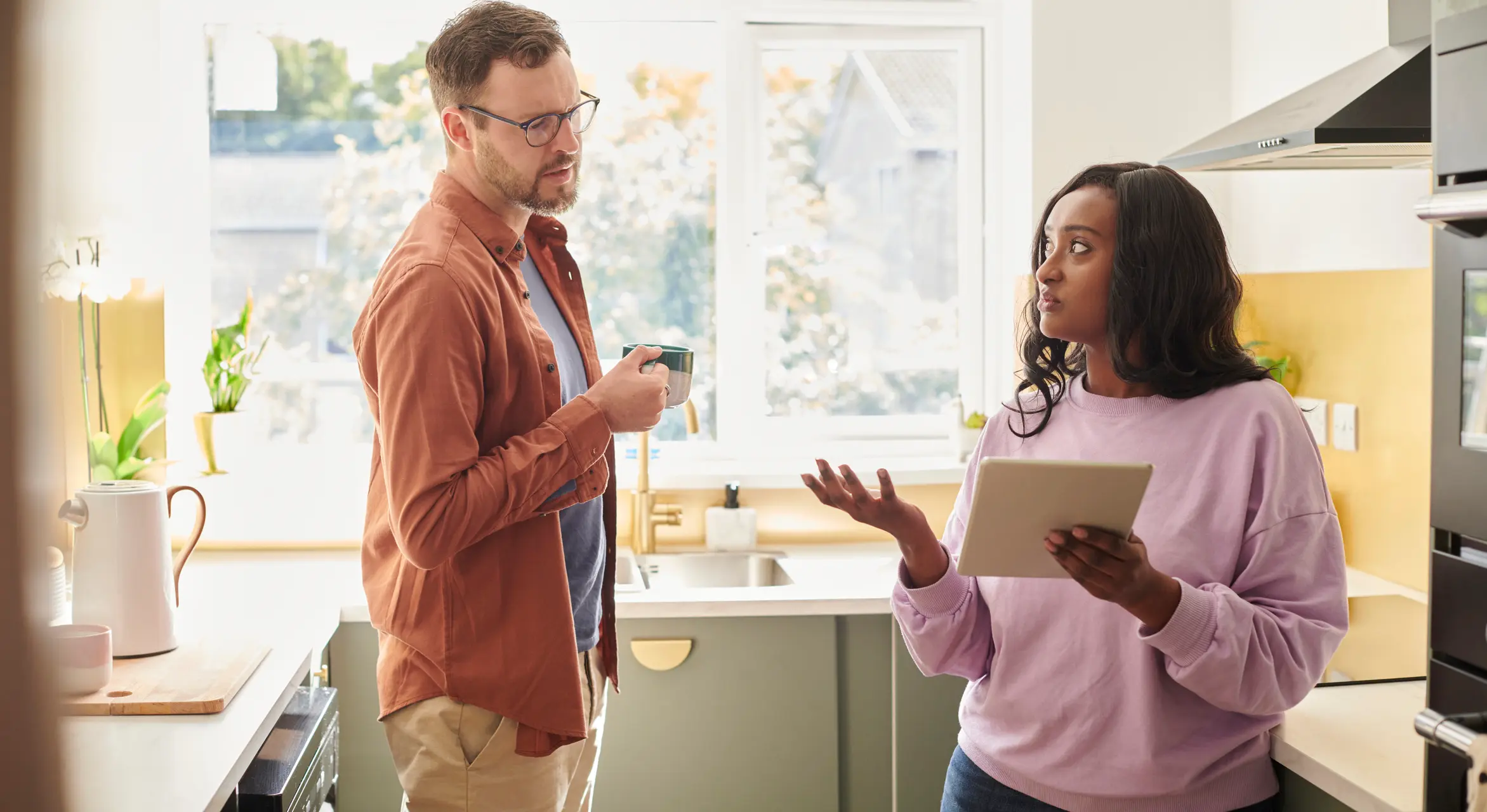 A young couple in their kitchen in discussion while the woman holds a tablet device up to her partner.